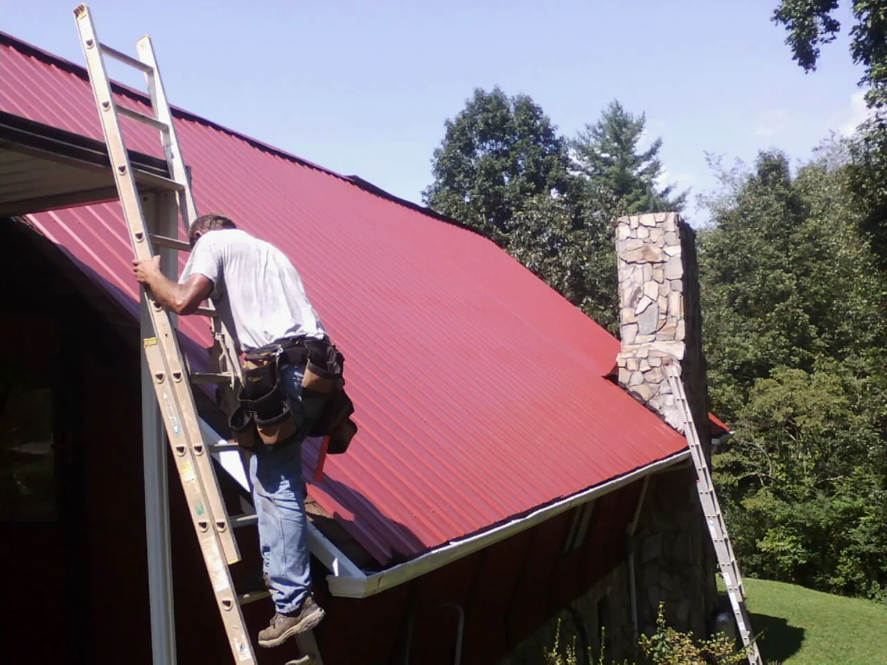 Roofer on a ladder beside a red metal roof and stone chimney — Soffit & Fascia Repair by Roofing Brothers in Luling